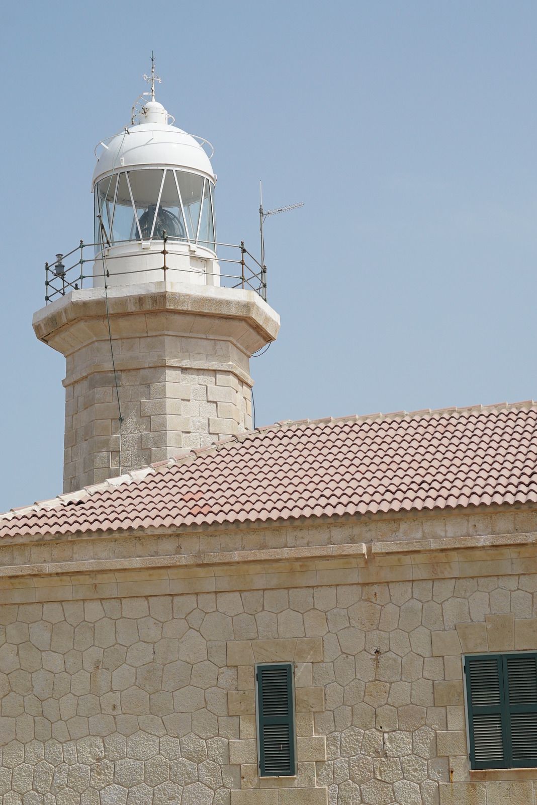 Stone lighthouse tower with a white lantern beside a tan stone building under a clear blue sky