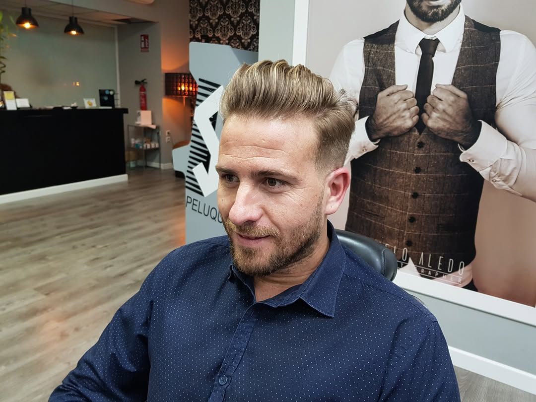 Un barbero le corta el pelo a un cliente en un estudio bien iluminado, con un reloj de pared al fondo.
