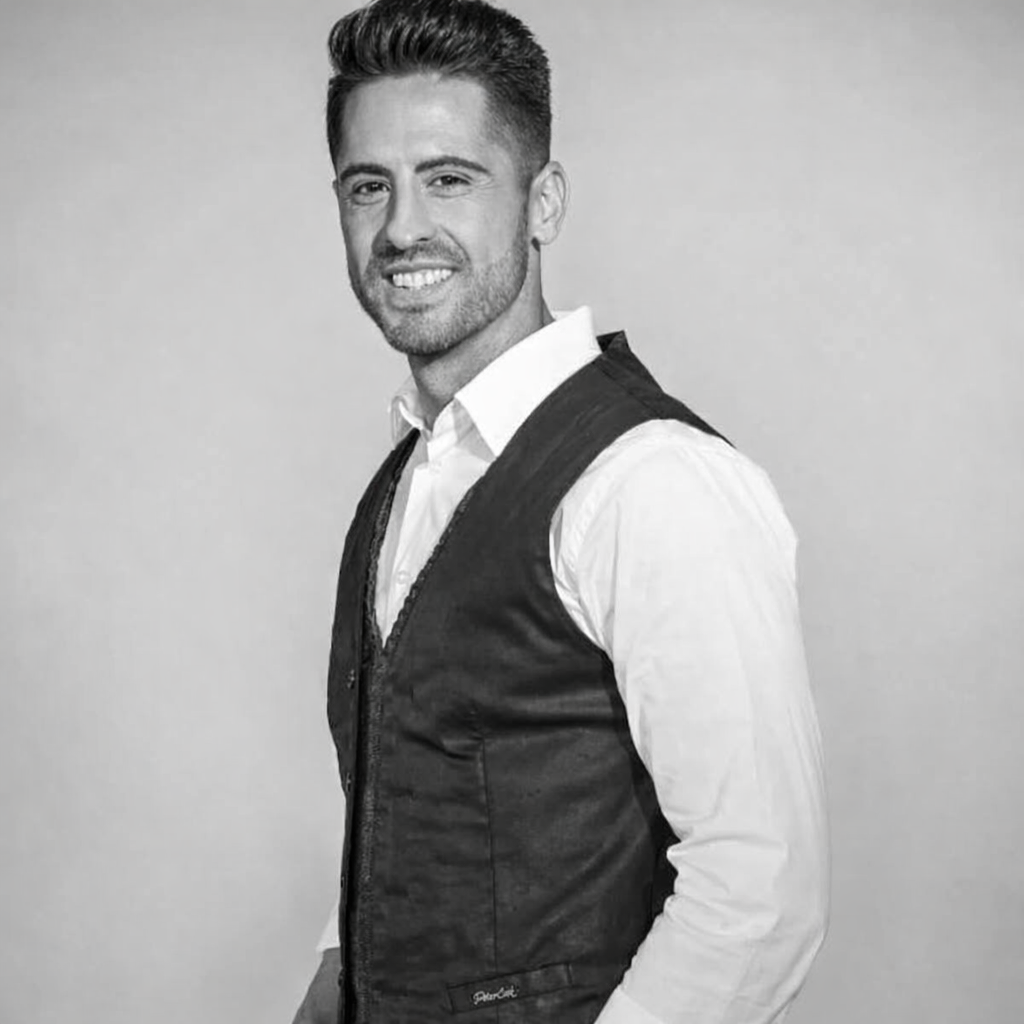 Una persona con una camisa blanca abotonada y un chaleco oscuro, sonriendo sobre un fondo gris neutro.