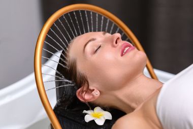 Un homme au visage recouvert d'une substance blanche est allongé sous un distributeur d'eau doré incurvé, sur fond sombre.