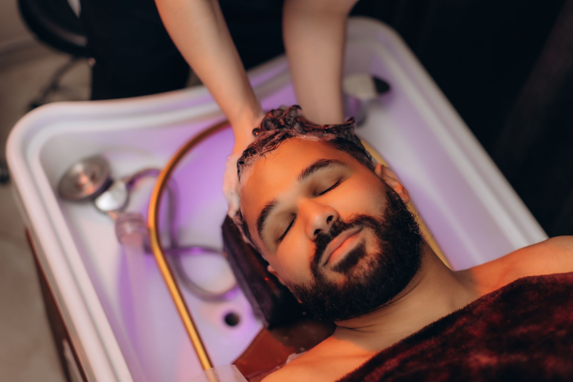 Un homme se fait laver les cheveux dans un salon de coiffure, les yeux fermés, la tête dans un lavabo.