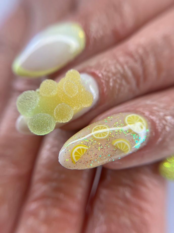 Gros plan sur une main ornée d'une manucure sur le thème du citron jaune, comprenant des motifs d'oursons en gélatine en 3D et de tranches de citron.