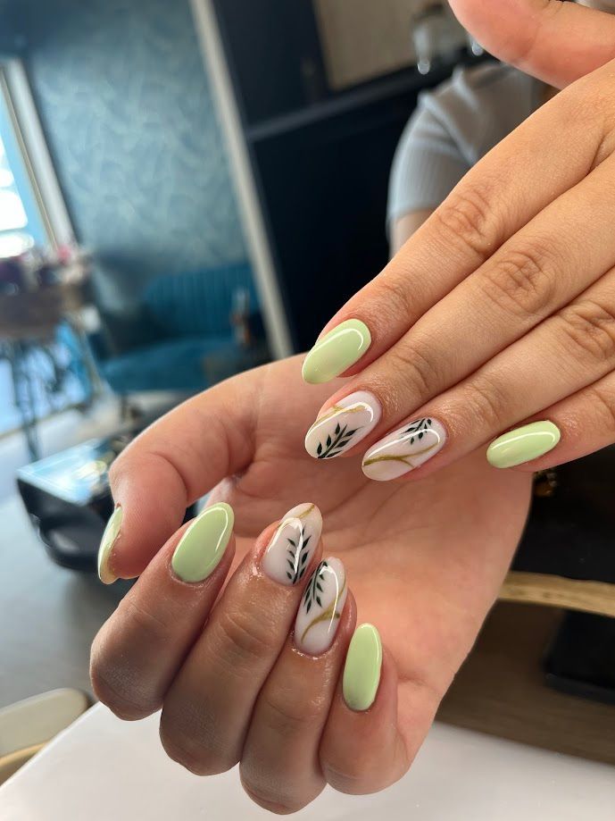 Des mains aux ongles en amande, vernis en vert clair et ornés de motifs de feuilles sur certains ongles.