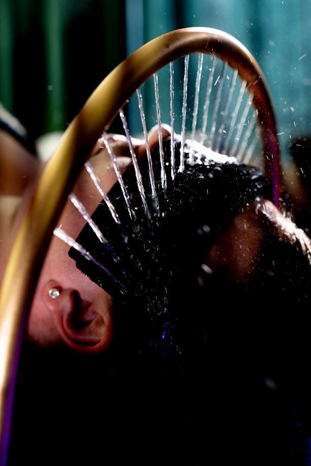 Une personne se cambre en arrière sous un cerceau qui l'arrose. Ses yeux sont dissimulés par un bandeau noir, des jets d'eau en cascade.