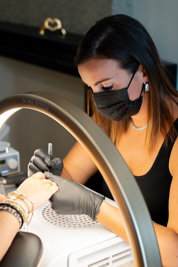 Femme portant un masque noir et des gants, appliquant du vernis à ongles sur la main d'une cliente, éclairée par un anneau lumineux.