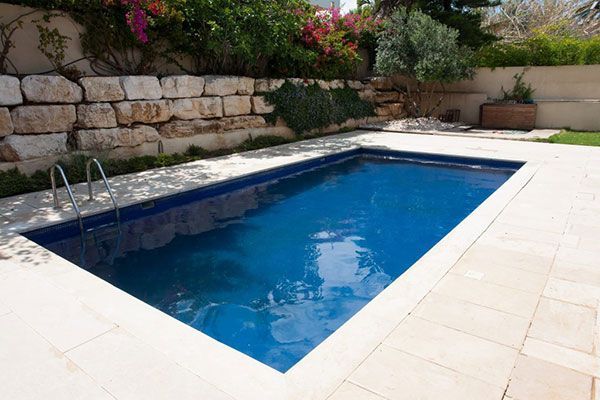 Piscine rectangulaire bleue jouxtant un mur en pierre et une terrasse carrelée et fleurie.