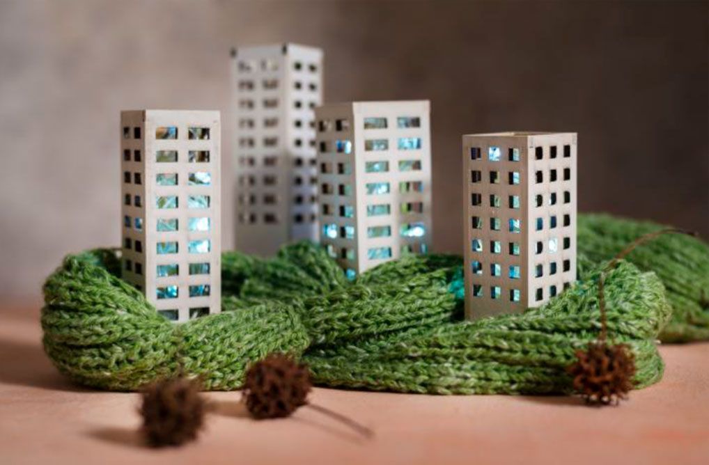 Edificios en miniatura de papel blanco con ventanas iluminadas reposan sobre una bufanda verde de punto. Dos piñas descansan delante.