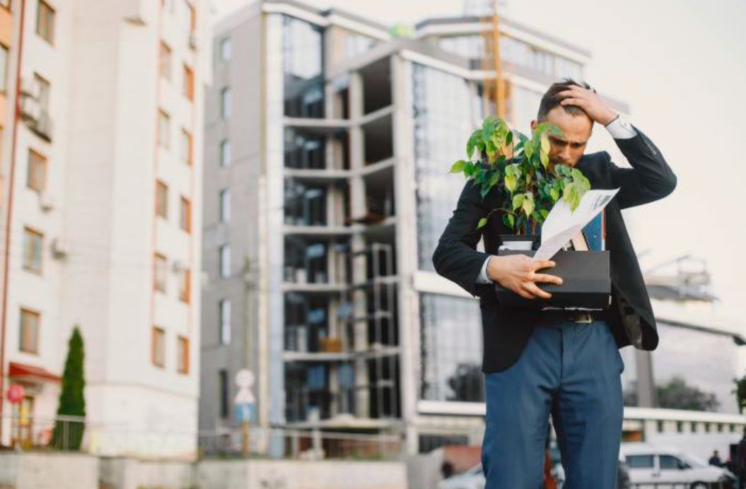 Hombre sale de un edificio de oficinas, cargando una caja con sus pertenencias, con aspecto estresado.