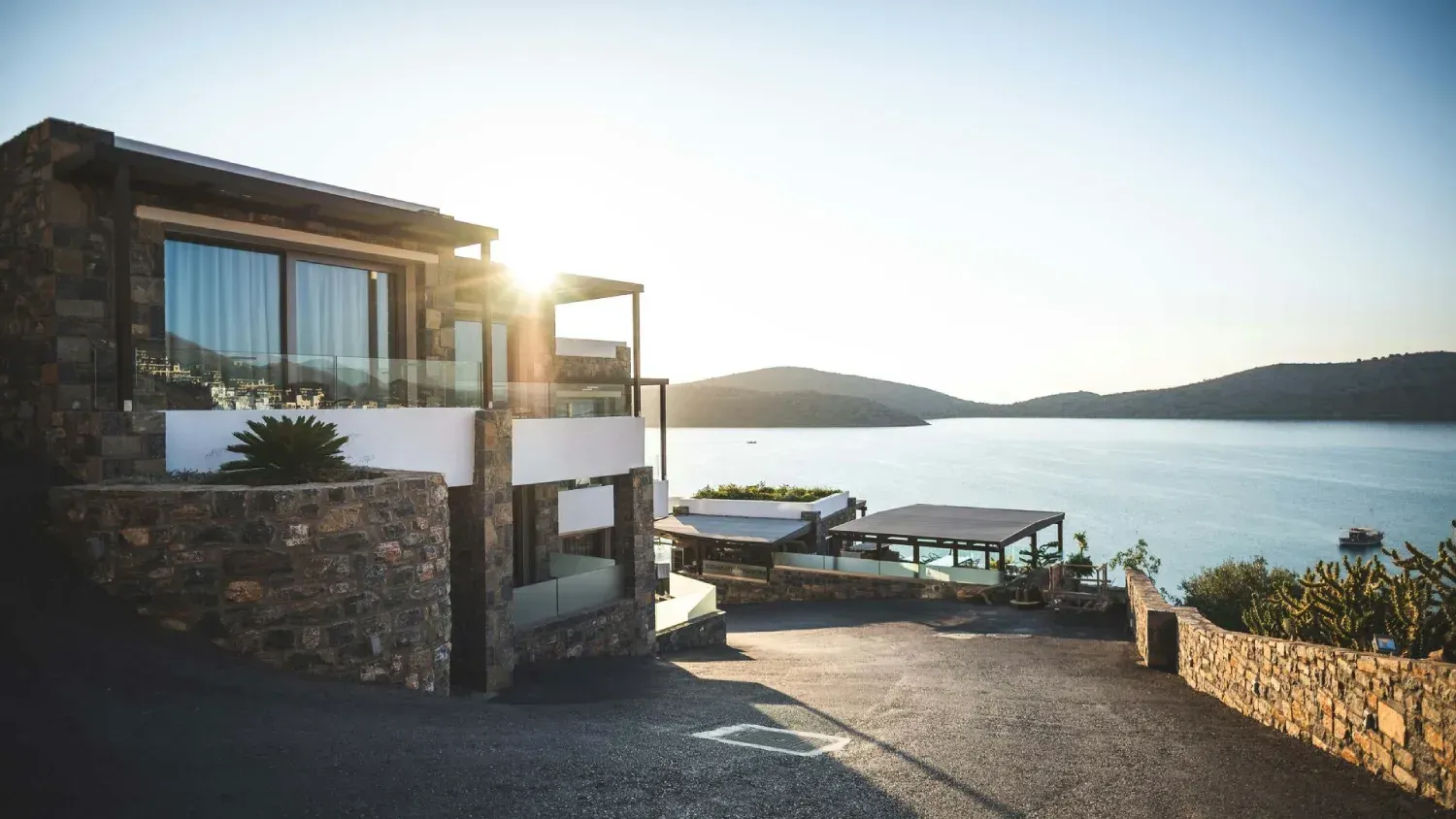 Edificio de piedra con balcón con vistas a la bahía al atardecer; un camino sinuoso conduce a un pequeño patio exterior.