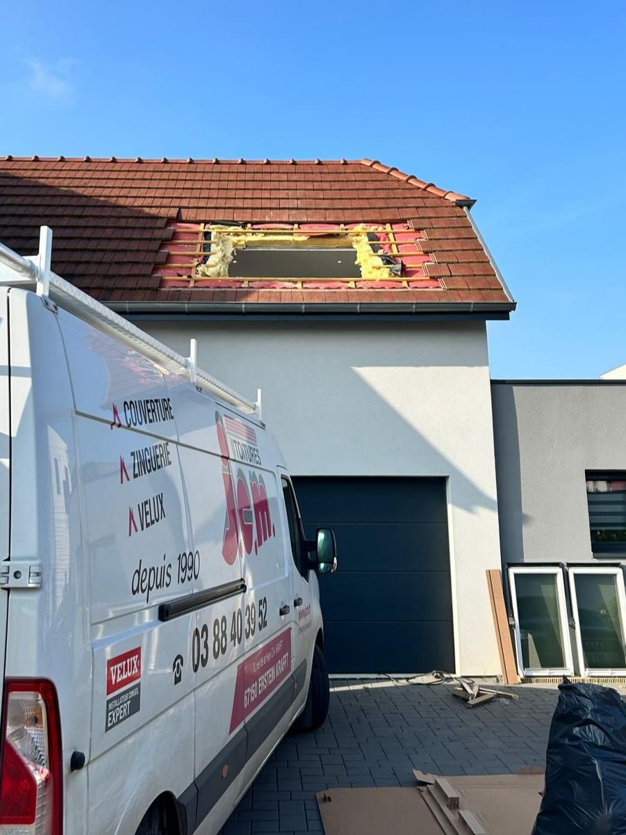 Une camionnette blanche avec un logo est garée devant une maison dont le toit est endommagé. Des fenêtres neuves se trouvent à proximité. Ciel bleu.