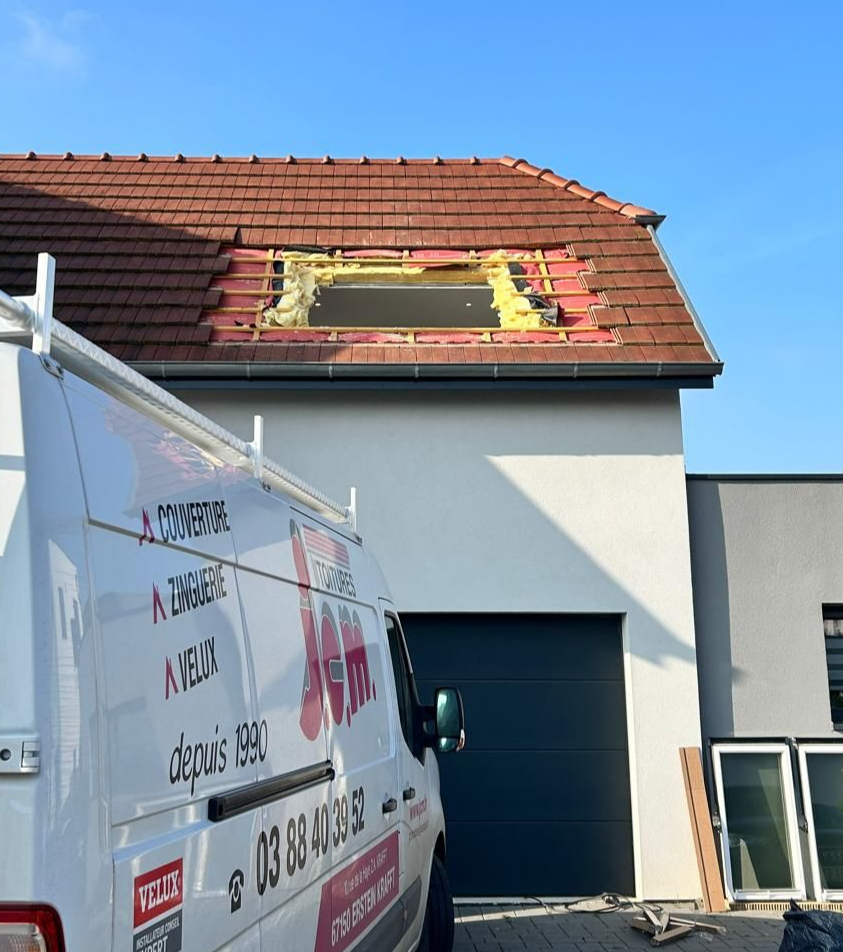 Une camionnette blanche est garée devant une maison où des travaux de réparation de toiture sont en cours, laissant apparaître les chevrons.
