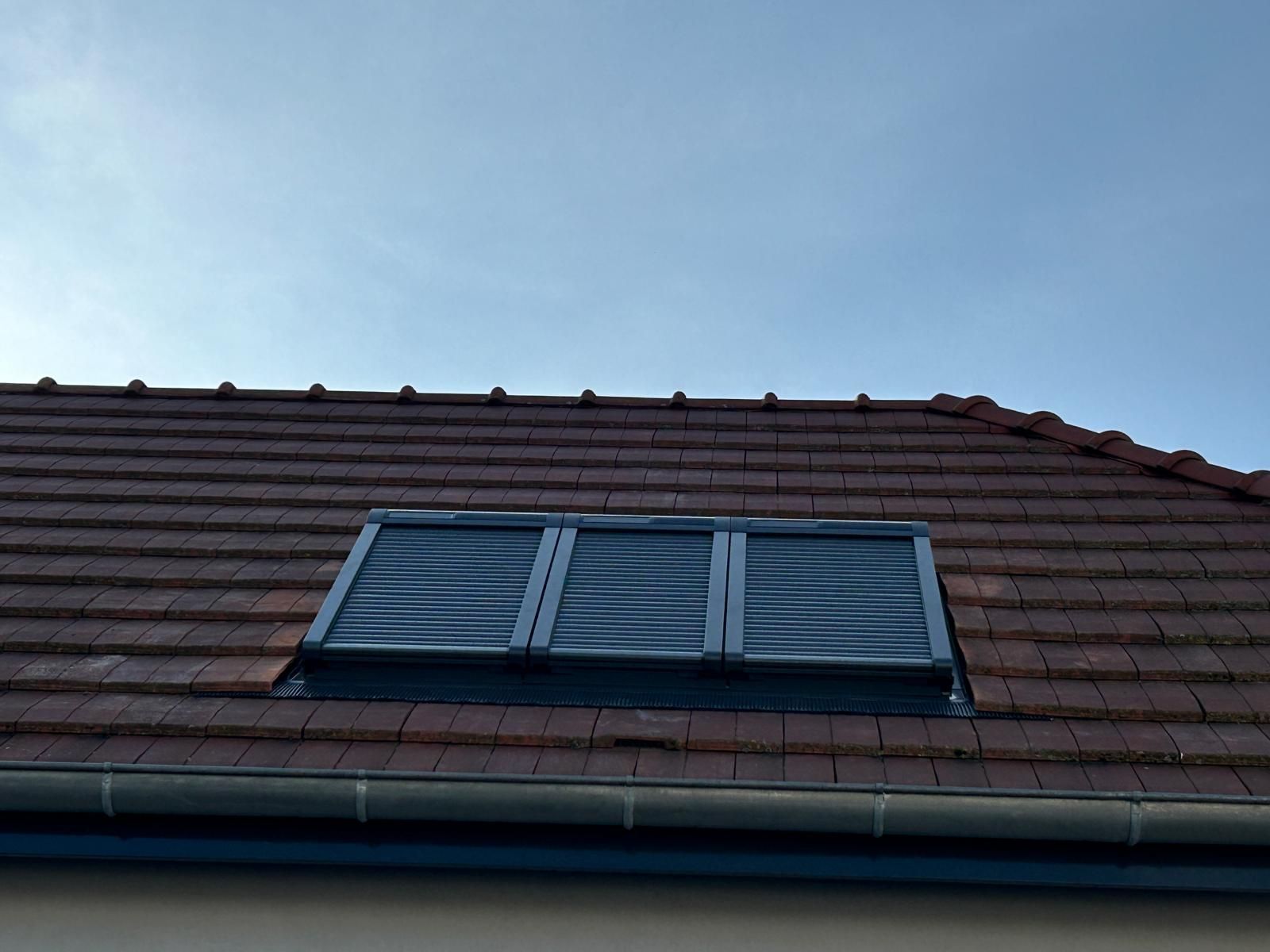 Toit en tuiles avec une lucarne aux volets gris fermés, sur fond de ciel bleu.