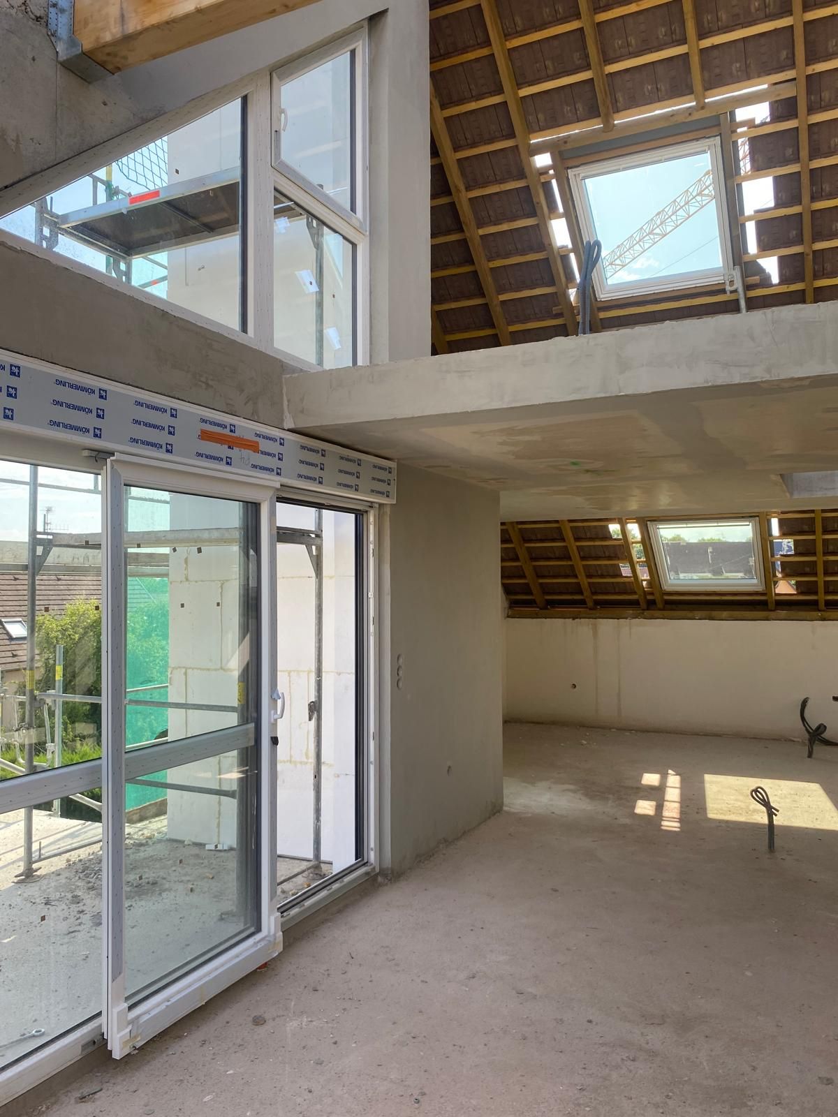 Intérieur d'un bâtiment neuf en construction ; grandes fenêtres, sols en béton, poutres apparentes au plafond et puits de lumière.