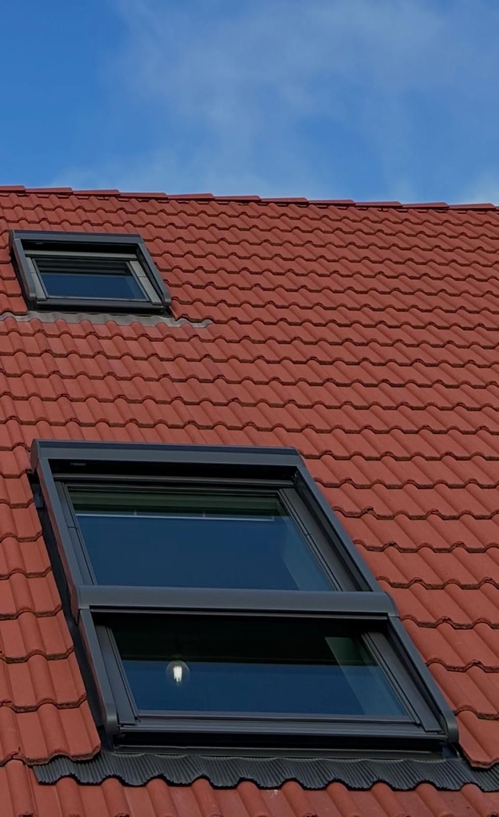 Toit de tuiles rouges percé de deux puits de lumière sur fond de ciel bleu, l'un ouvert, l'autre fermé.