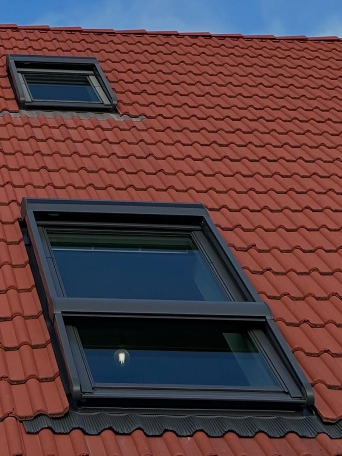 Toit de tuiles rouges avec deux puits de lumière ; l'un ouvert, l'autre fermé, sur fond de ciel bleu.