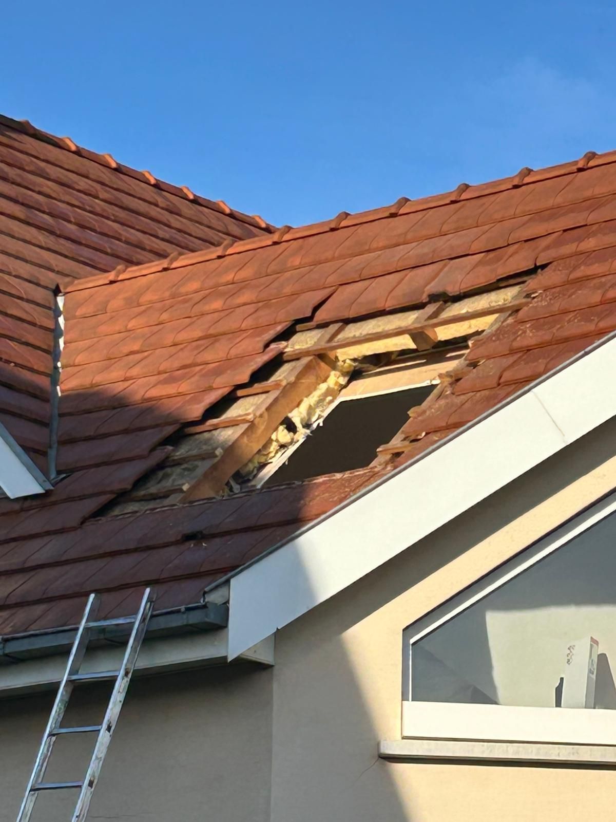 Toiture en tuiles rouges endommagée avec un trou, charpente en bois apparente et une échelle.
