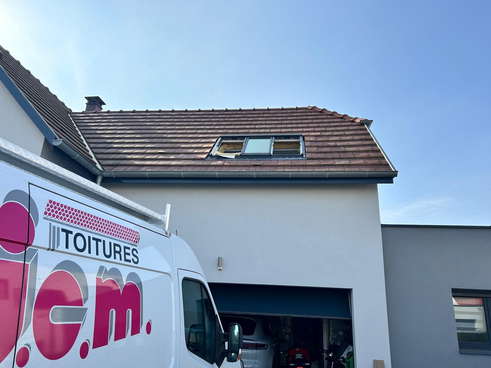 Une camionnette blanche est garée devant une maison ; on aperçoit le toit avec une fenêtre de toit.