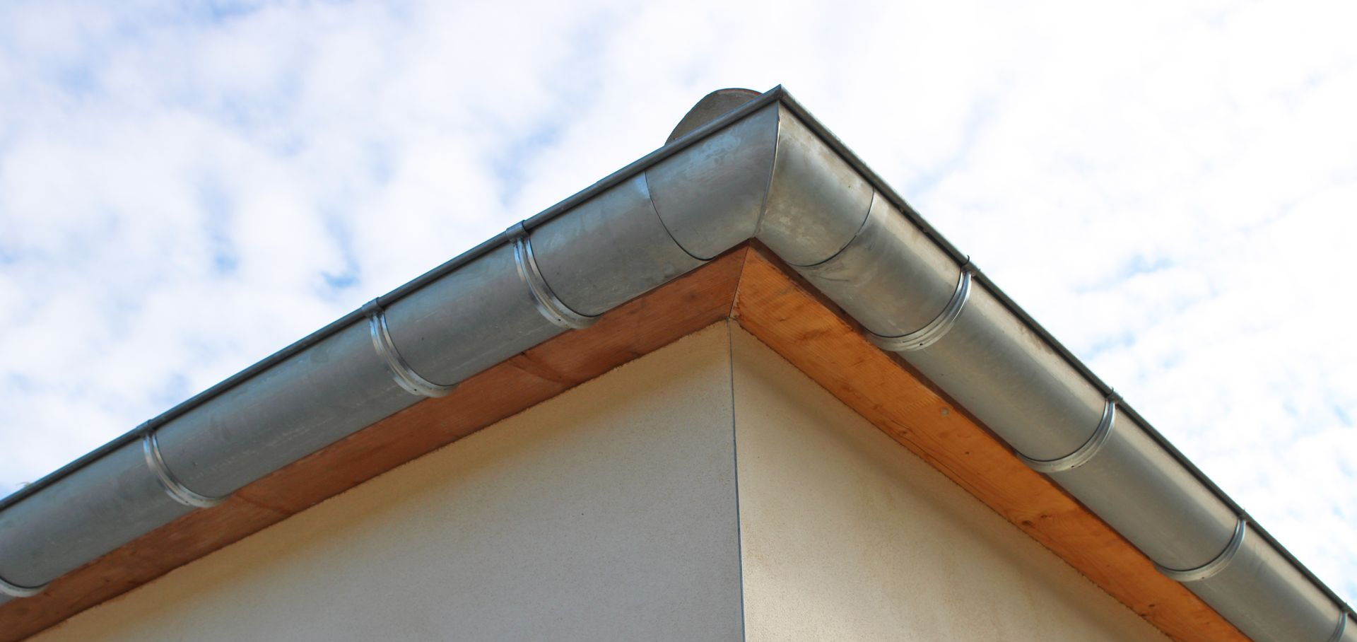 Toiture en tuiles de terre cuite, gouttière grise et descente pluviale sur un mur en stuc beige, le tout se détachant sur un ciel bleu nuageux.