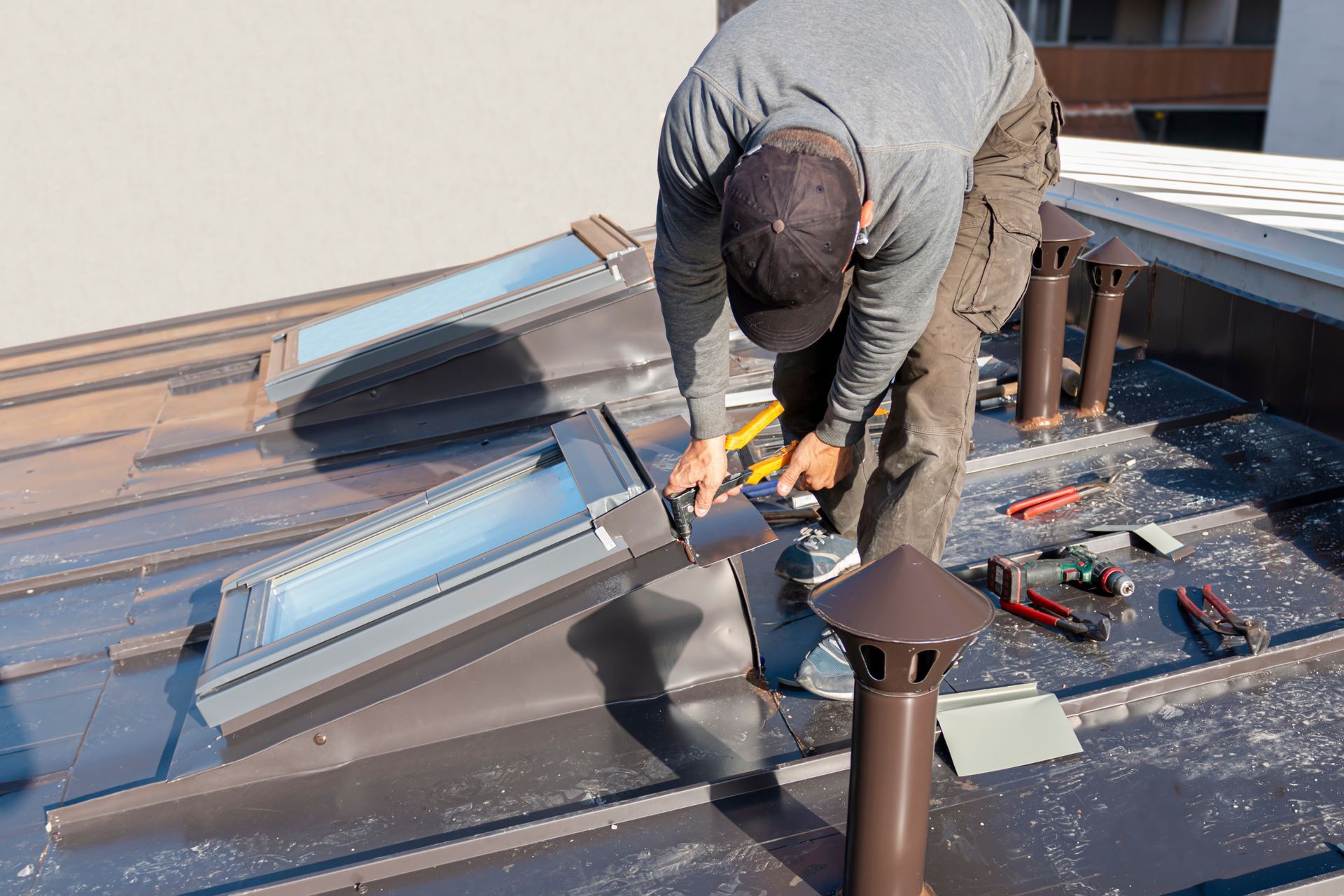 Un couvreur installe des solins autour d'un puits de lumière sur un toit en métal. On aperçoit des tuyaux bruns, des outils et la lumière du soleil.