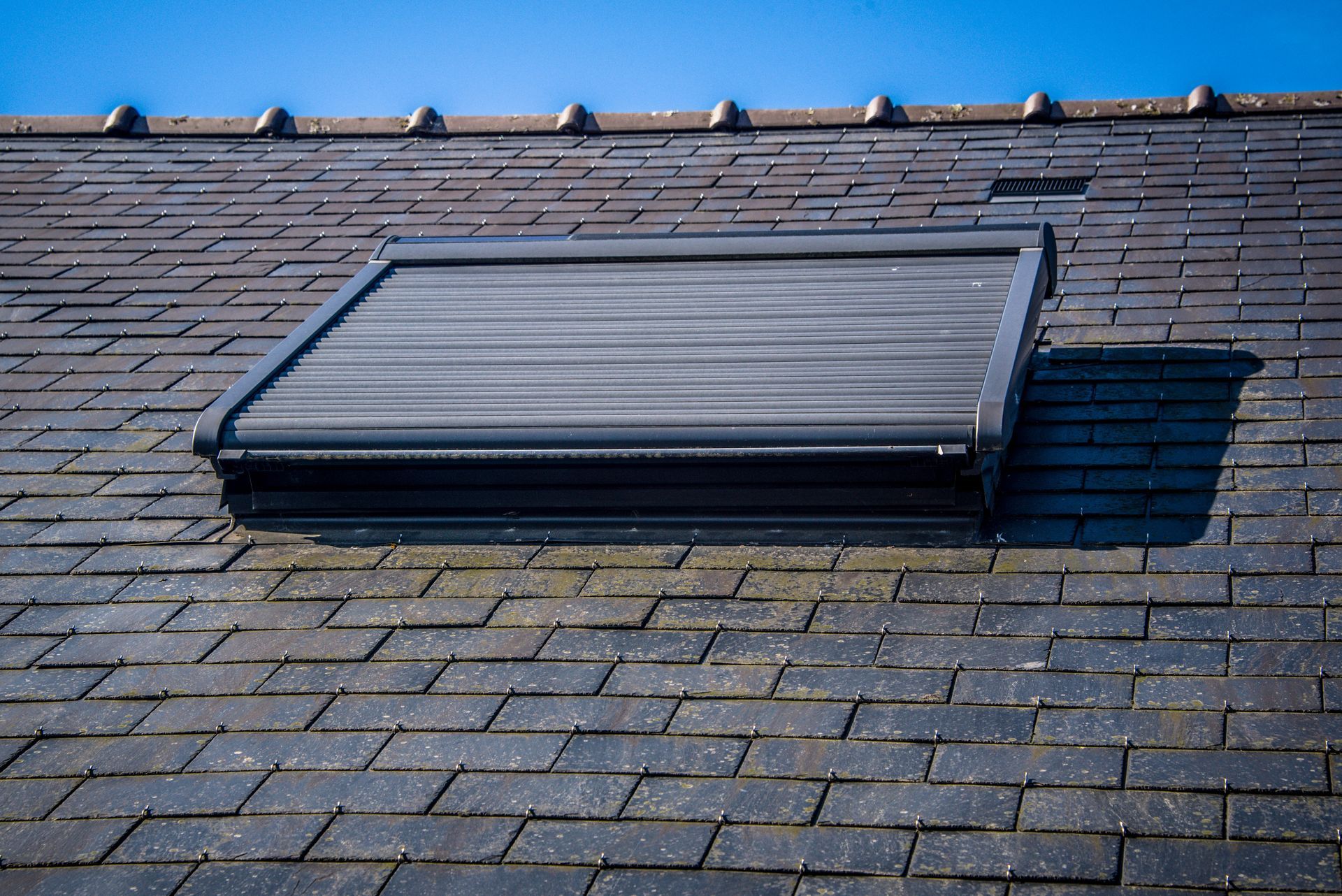 Toit de tuiles noires avec une lucarne ouverte sur un ciel bleu.