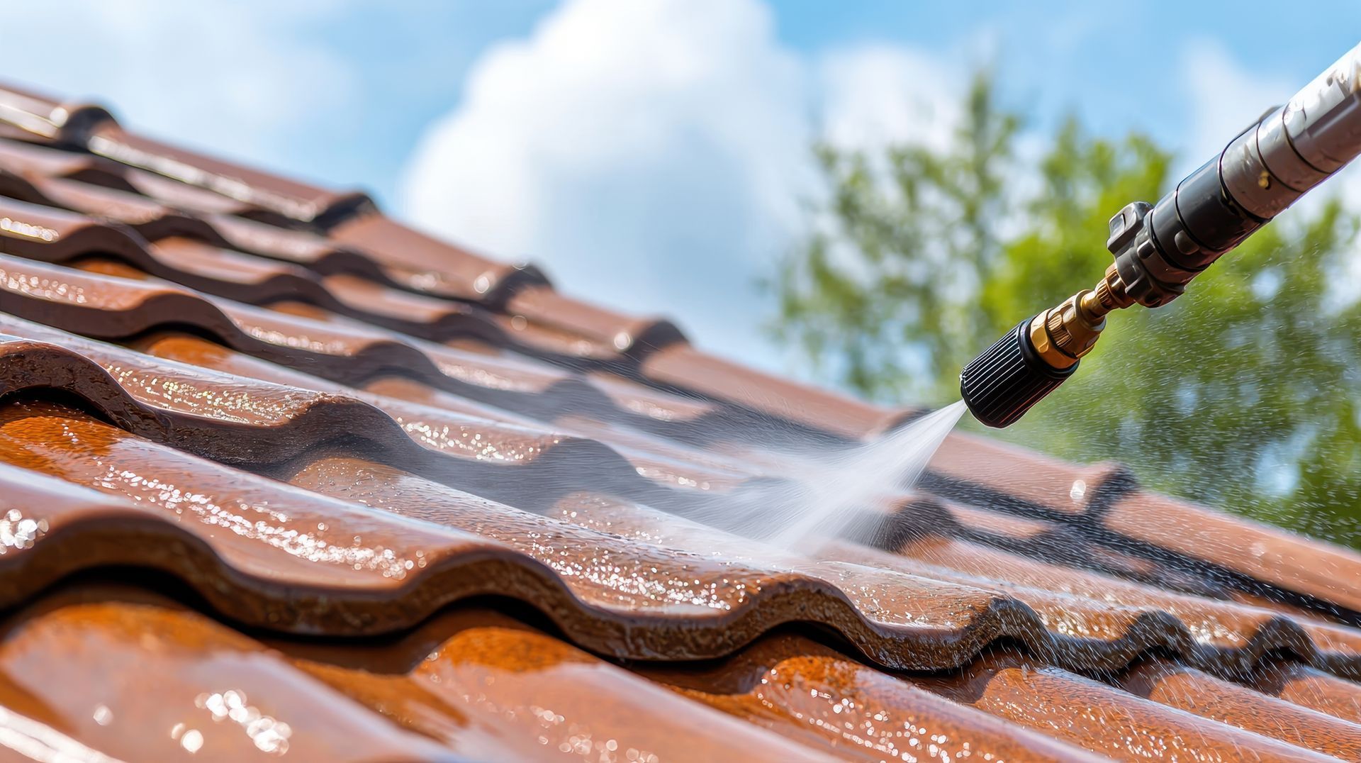 Nettoyeur à haute pression arrosant un toit de tuiles brunes ; ciel bleu et arbres en arrière-plan.