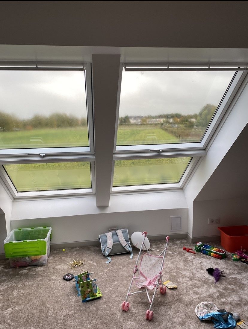 Chambre avec fenêtres en biais donnant sur un champ verdoyant. Jouets éparpillés sur le sol recouvert de moquette.