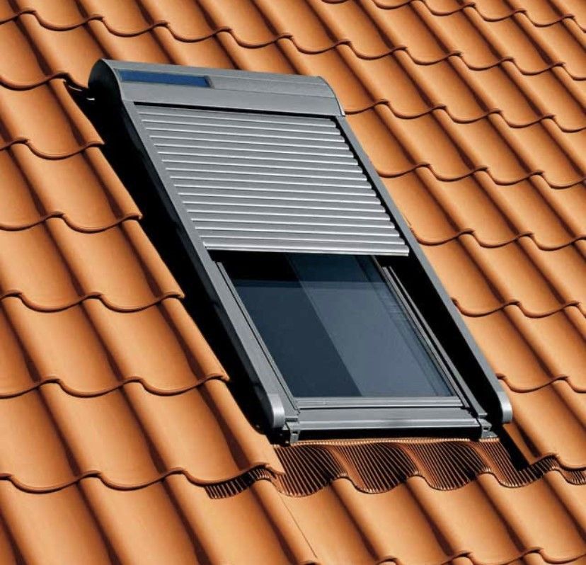 Puits de lumière sur un toit de tuiles avec un volet roulant gris fermé.
