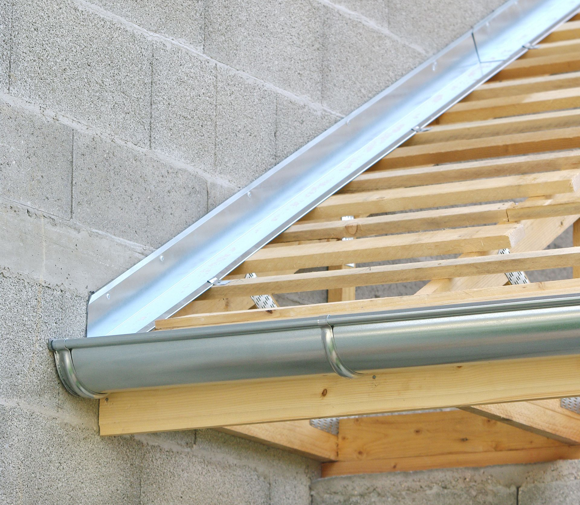 Gouttières et solins gris installés sur un toit à chevrons en bois et un mur en blocs de béton.