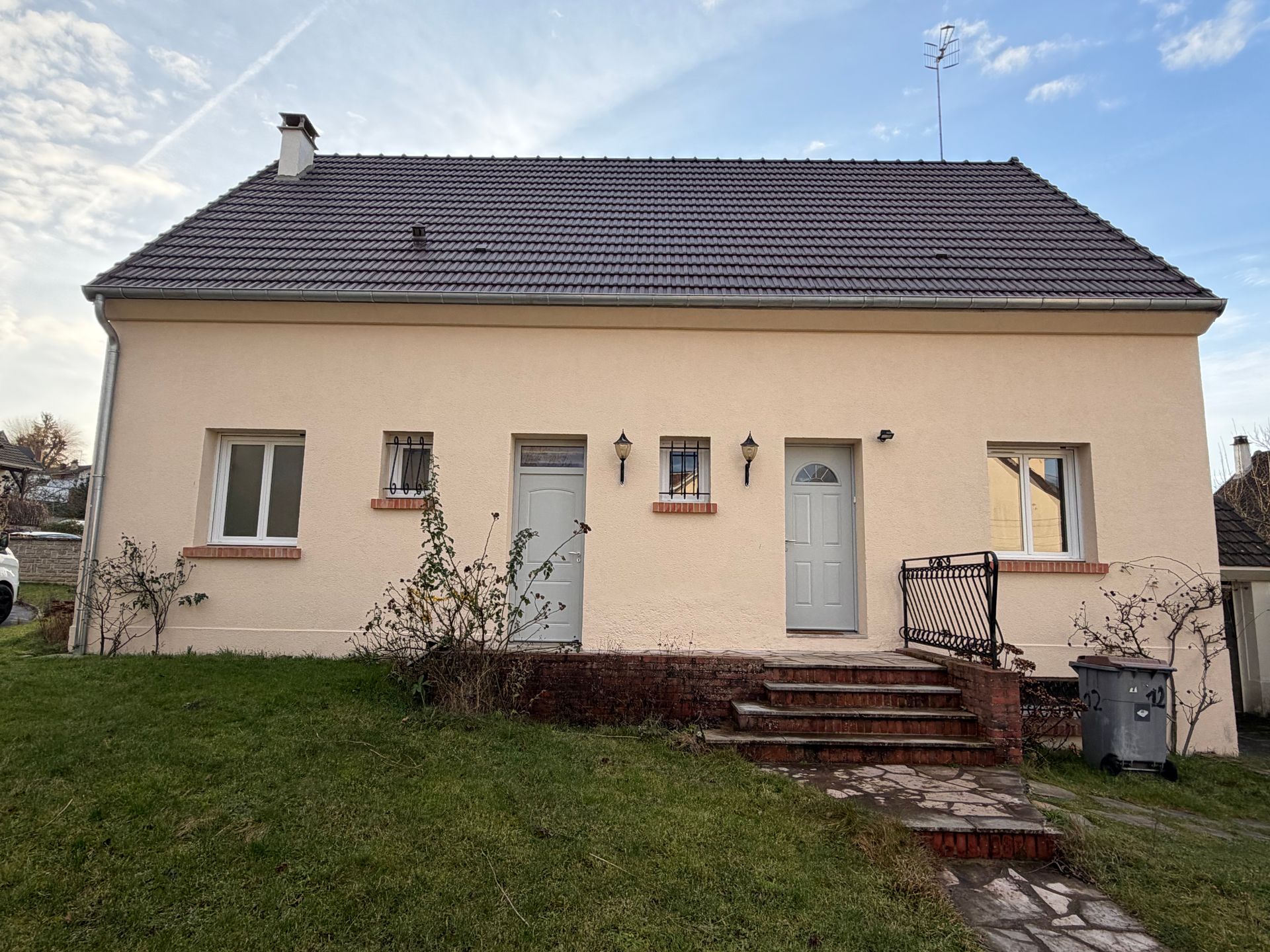 Maison beige avec toit gris, deux portes d'entrée et une petite cour avant avec des marches.