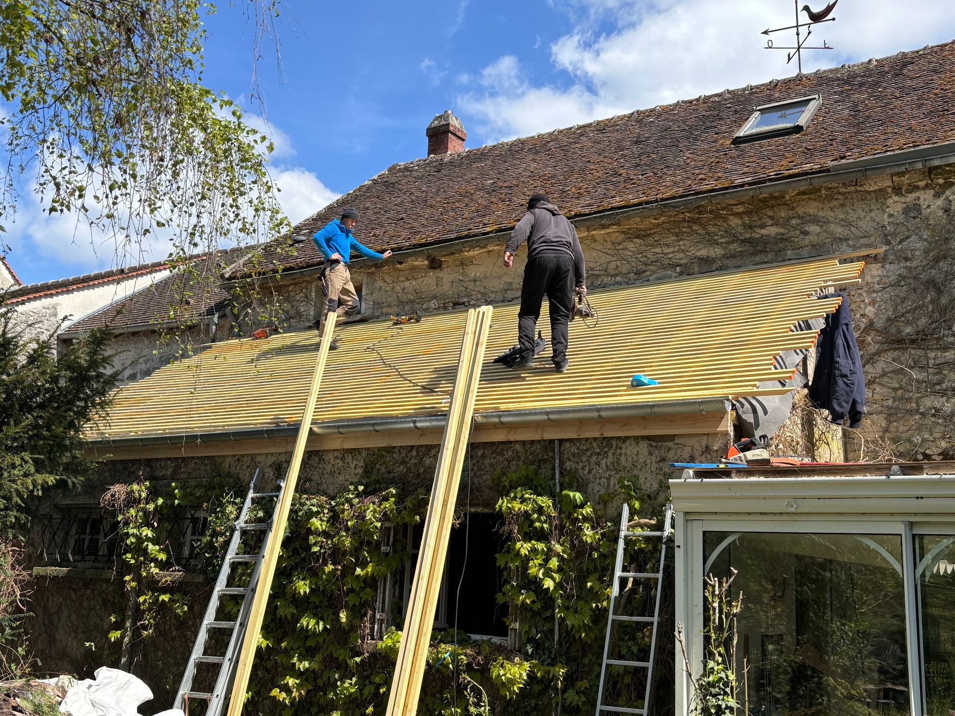 Deux personnes sur des échelles travaillent sur un toit recouvert d'une couverture à motifs tourbillonnants, bâtiment en pierre, journée ensoleillée.