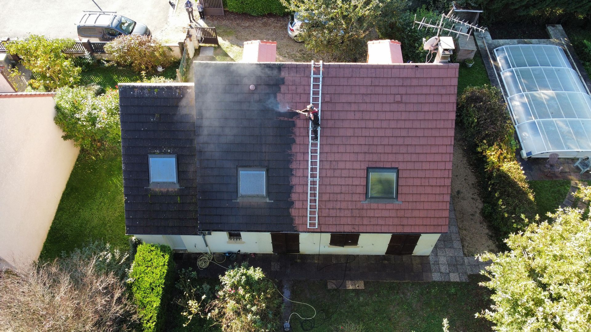 Une maison dont le toit est en cours de nettoyage ; un côté est sombre, l'autre rouge, avec une échelle et une personne dessus.