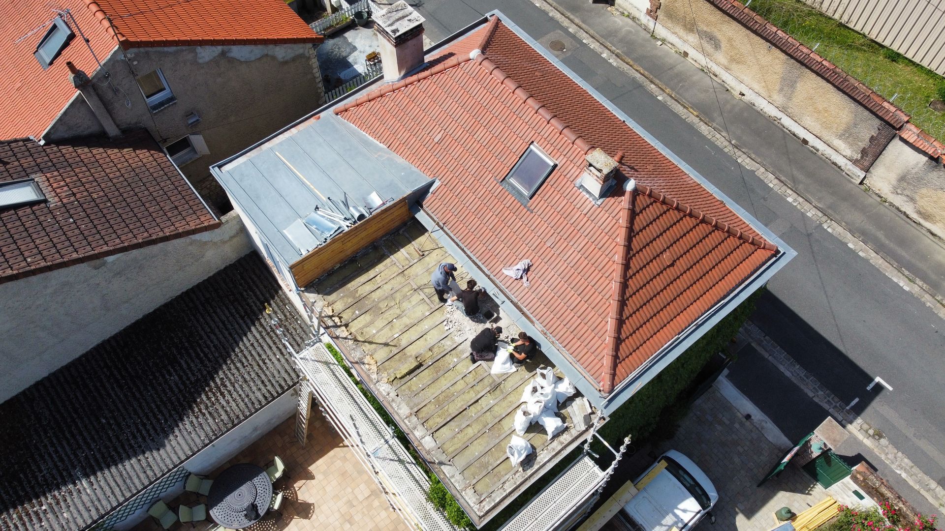 Vue aérienne d'un toit de tuiles rouges avec une section plate recouverte d'un toit argenté, sur laquelle figurent plusieurs personnages, et des bâtiments environnants.