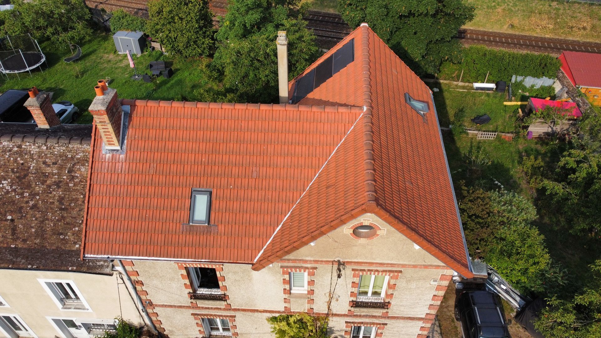 Vue aérienne d'une maison au toit de tuiles rouges, avec deux cheminées et une façade en briques beiges.