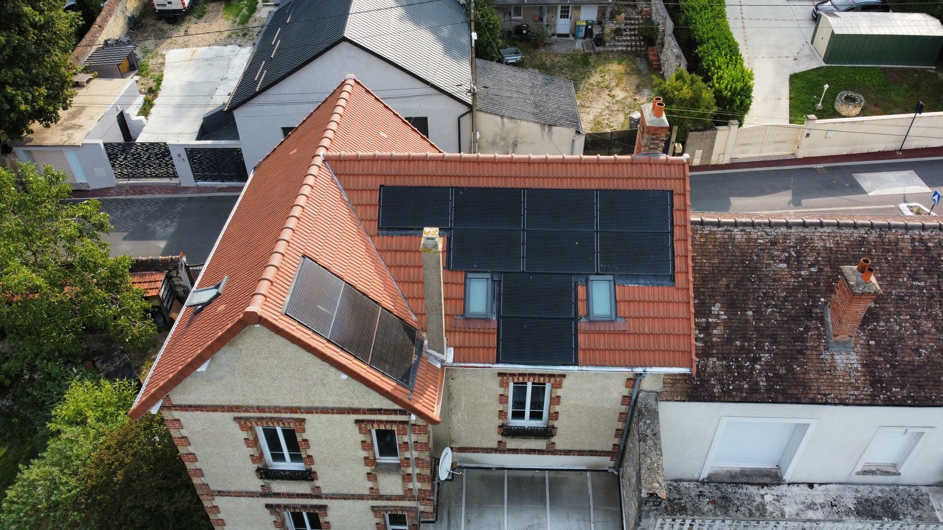 Vue aérienne d'une maison avec un toit de tuiles rouges, des panneaux solaires, des fenêtres et une cheminée.