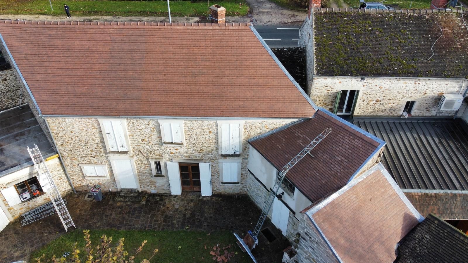 Maison en pierre avec un toit de tuiles brunes, un autre toit à l'avant et des bâtiments environnants.