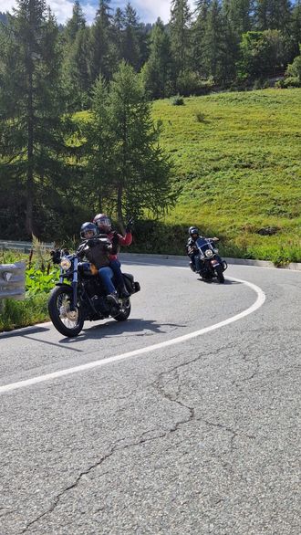 Deux motocyclistes roulent sur une route de montagne asphaltée en virage, bordée d'arbres et de collines verdoyantes.
