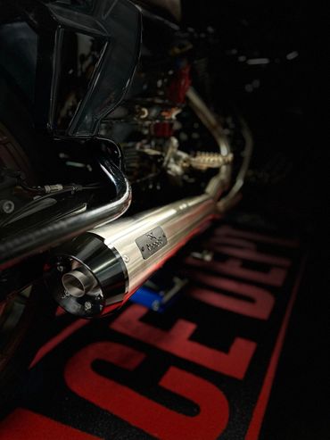 Gros plan sur un pot d'échappement argenté de moto avec un embout noir, placé au-dessus d'un tapis de sol noir et rouge.