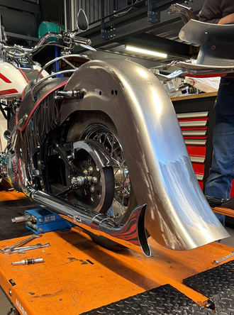 Un grand garde-boue de moto métallique incurvé, monté sur un cadre, exposé dans un atelier sur un pont élévateur orange.