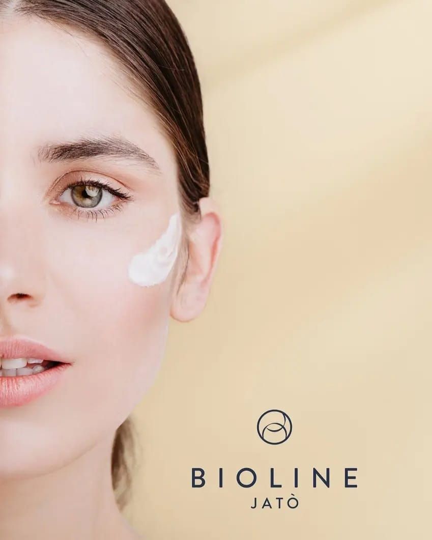 Primer plano de un rostro con crema en la mejilla sobre un fondo beige, junto al logotipo y el nombre de la marca Bioline Jató.