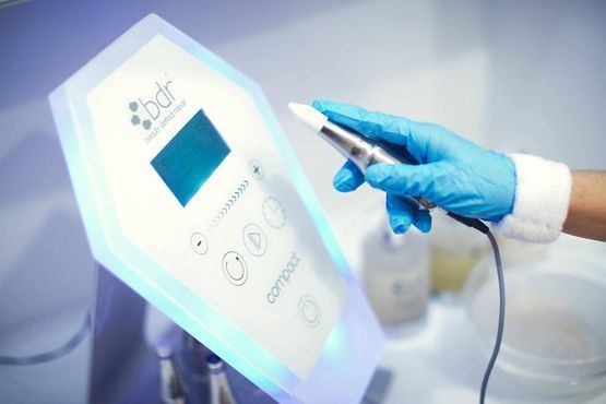 Una persona con guantes azules sostiene un dispositivo profesional para el cuidado de la piel en un entorno médico o de spa.