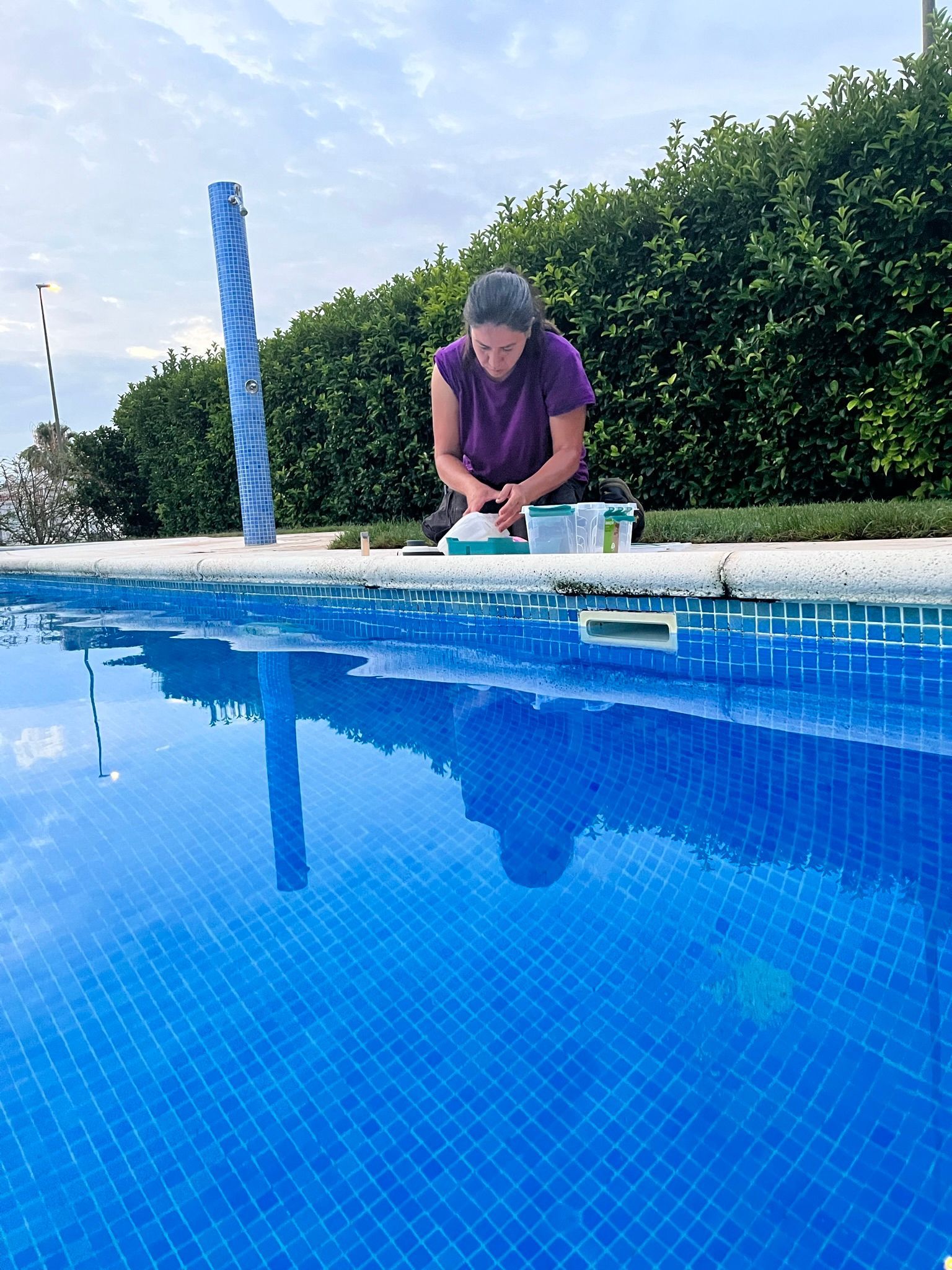 Persona arrodillada junto a una piscina azul, limpiando o trabajando cerca del borde del agua.