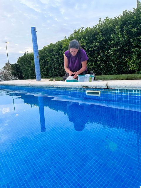 Persona arrodillada junto a una piscina azul, limpiando o trabajando cerca del borde del agua.