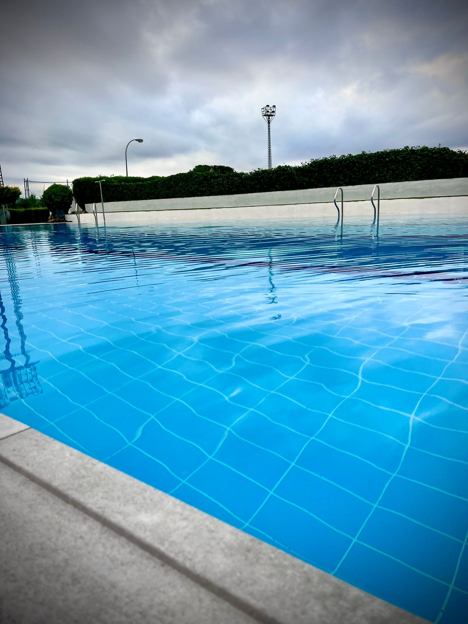 Piscina de color azul brillante con agua ondulante bajo un cielo nublado, zona de descanso blanca y vacía al fondo.