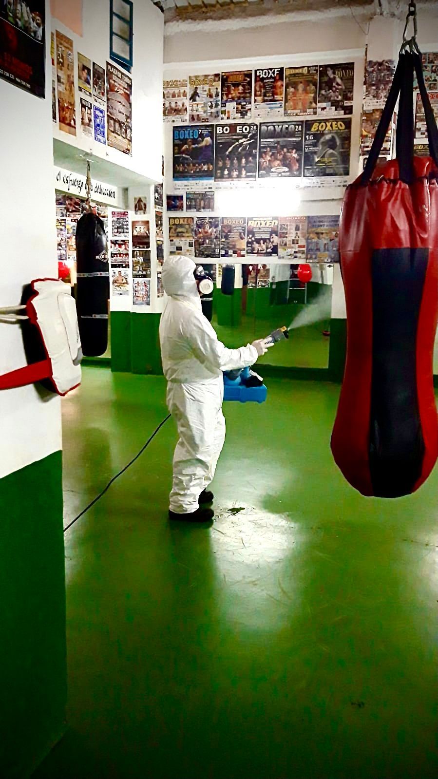 Una persona con un traje protector blanco se encuentra en un gimnasio de boxeo junto a un saco de boxeo rojo sobre un suelo verde.