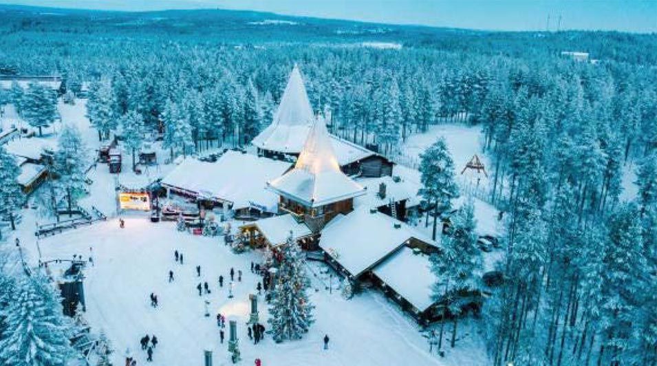 Pueblo nevado con edificios iluminados y árboles altos. La gente pasea por una zona decorada en el paisaje invernal.