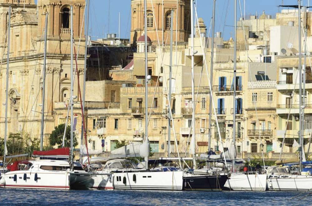 Veleros atracados en un puerto con un fondo de edificios de colores claros bajo un cielo azul.