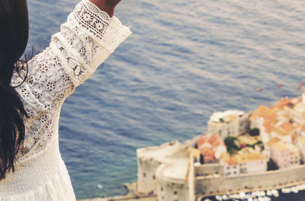 Una mujer con mangas de encaje blanco levanta el brazo hacia un pueblo costero. El océano azul está de fondo.