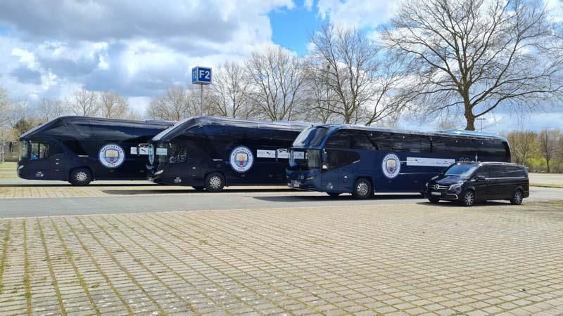 Drei blaue Busse und ein schwarzer Lieferwagen stehen auf einem Parkplatz.