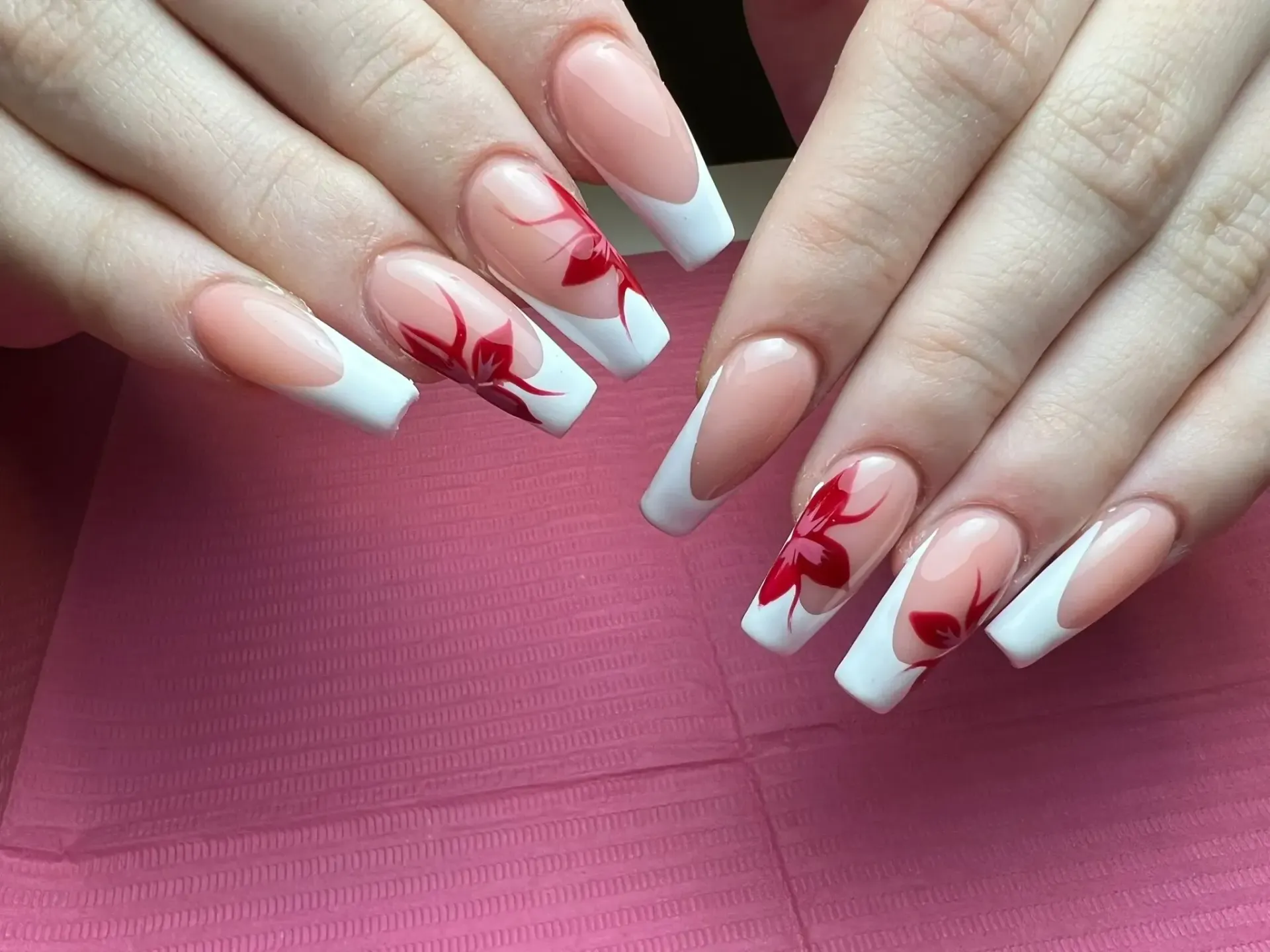 Manos con uñas de punta francesa, cada una con un diseño de flores rojas sobre una superficie rosa.