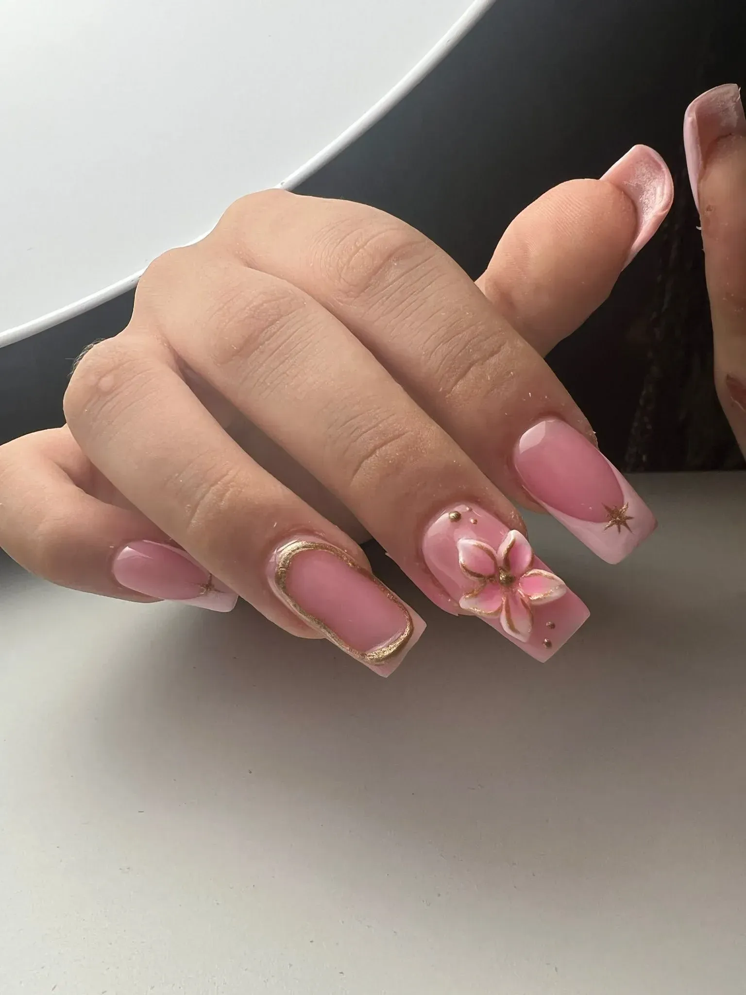Mano con uñas cuadradas rosas, puntas francesas y detalles dorados. Una uña tiene un diseño floral en 3D.